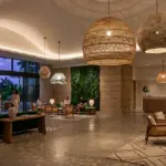 A panoramic view of a luxury hotel lobby with a biophilic design, featuring large organic-textured pendant lights, a green living wall, and a gold RST logo on a wood-paneled wall behind the reception desk.