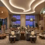 A wide-angle panoramic view of a modern minimalist hotel lobby at dusk, featuring low-profile grey sofas, woven and cane armchairs, and organic-textured pendant lights, with a gold RST logo prominently displayed on a textured stone wall behind the reception desk.