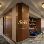 A panoramic view of a luxury hotel corridor featuring RST solid brass and resin flush mount ceiling lights, with a gold RST logo prominently displayed on a walnut wood pillar.